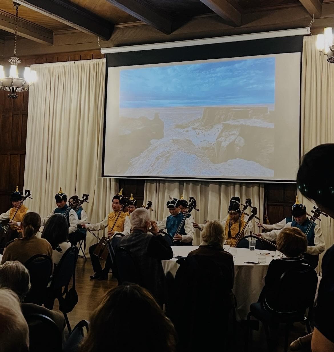Ajnai Ensemble playing morin khuur for a seated audience, with a steppe landscape projected behind them.