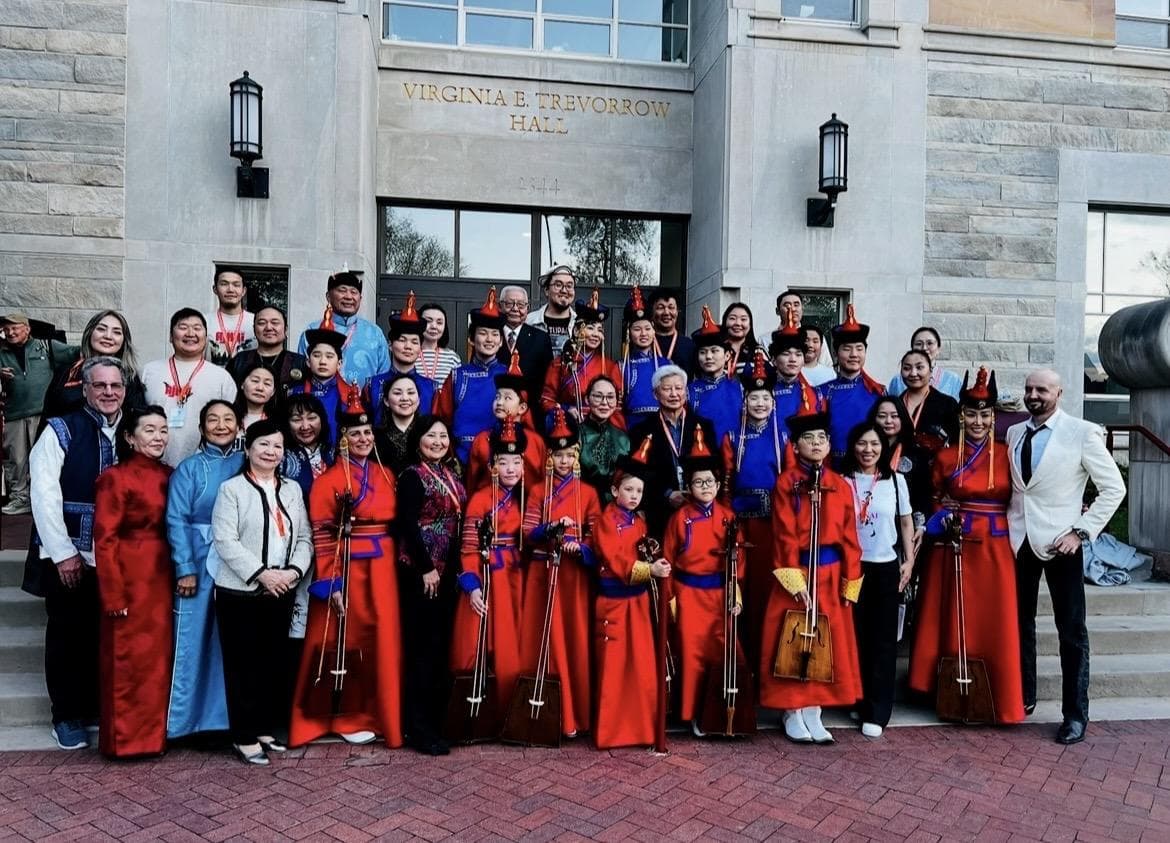 A large ensemble in red and blue deels with morin khuur, posed on the steps of Virginia E. Trevorrow Hall.