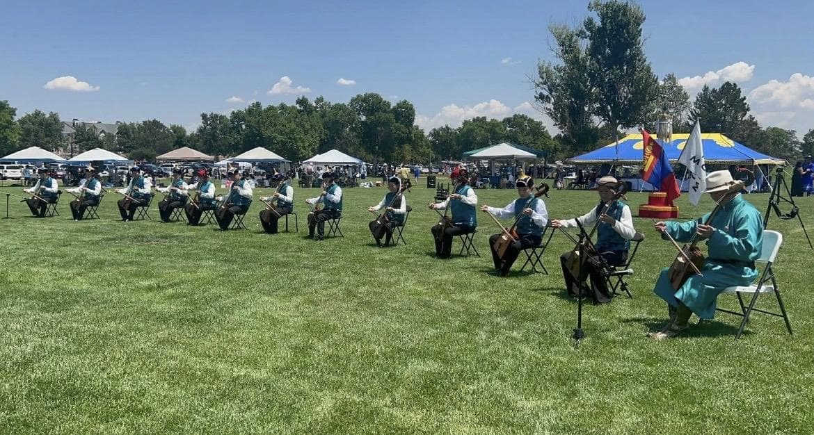 Musicians in teal vests playing morin khuur in a long line on a festival field, with a Mongolian flag nearby.