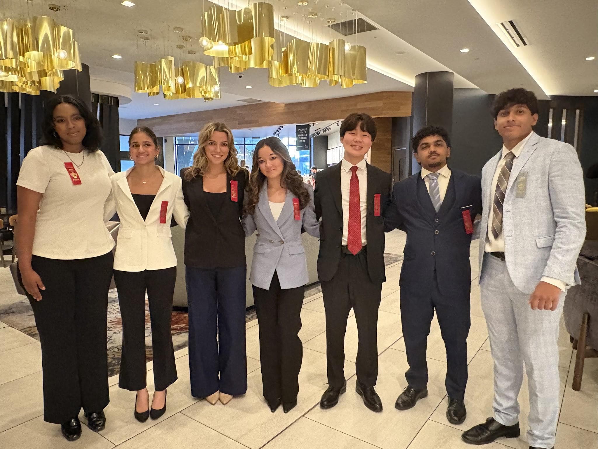 Seven competitors with red ribbons in a hotel lobby. Eric stands center-right in a black suit and red tie.