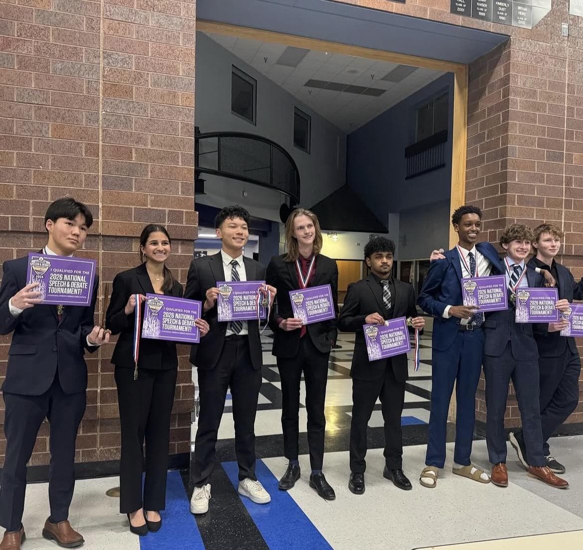Eric, at the far left in a navy suit and red tie, with other Colorado qualifiers holding signs for the 2026 National Speech & Debate Tournament.