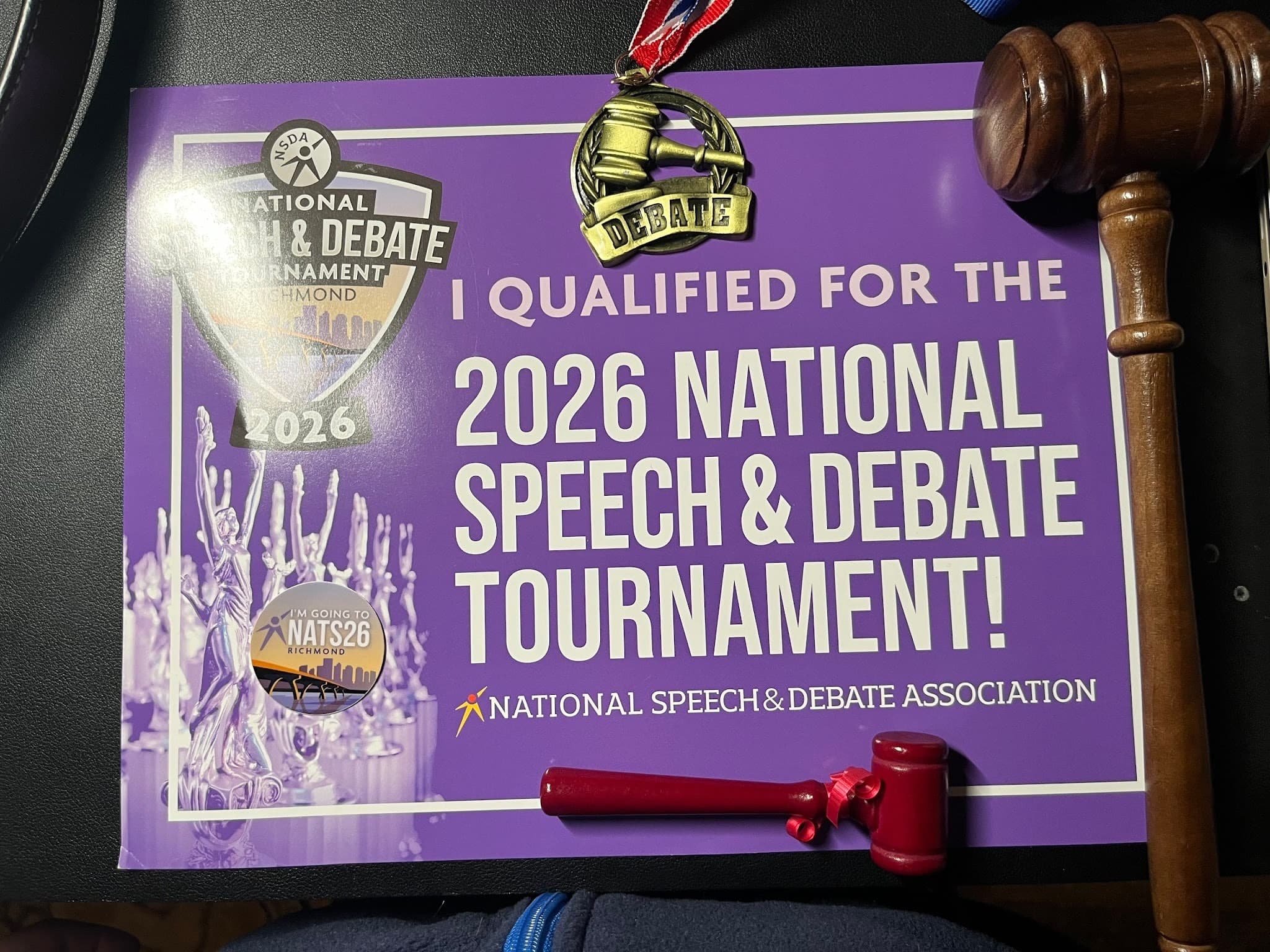 A 2026 National Speech & Debate qualifier sign with a debate medal, a small gavel, and a full-size gavel.