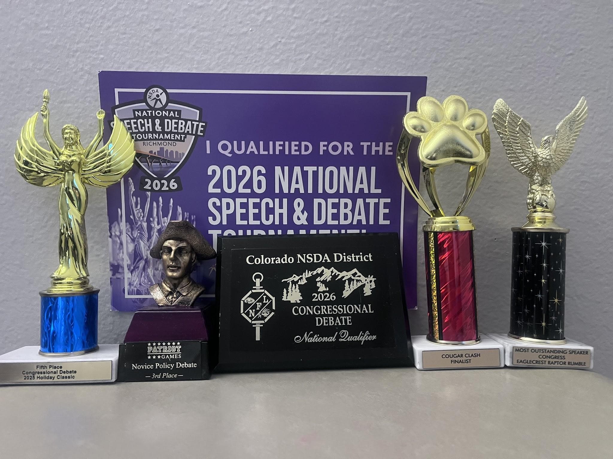 Debate trophies and a plaque in front of a 2026 National Speech & Debate qualifier sign, including Holiday Classic, Patriot Games, Cougar Clash, and Eaglecrest Raptor Rumble.